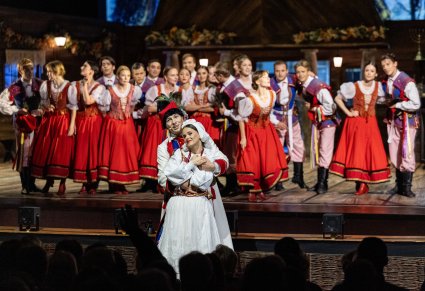 Na pierwszym planie przytulająca się Para Młoda w kostiumach krakowskich, w tle na scenie grupa Artystów w kostiumach ludowych