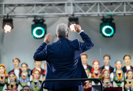 Zbliżenie od tyłu na dyrygującego Dyrektora zespołu, w tle widoczna część chóru w kostiumach łowickich