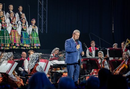 Na pierwszym planie Dyrektor, Dyrygent przemawiający do mikrofonu, widoczna część Orkiestry w kostiumach wilanowskich oraz część Artystów w kostiumach łowickich