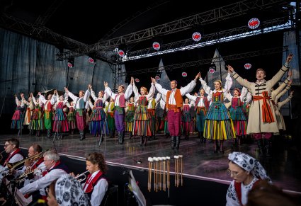 Artyści trzymający się za uniesione dłonie na scenie w kostiumach opoczyńskich a na dole część orkiestry