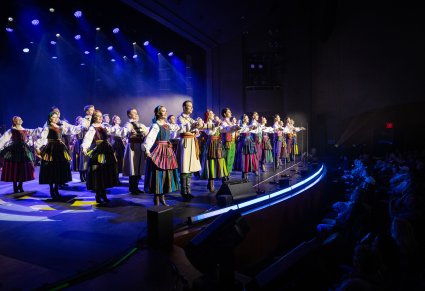 Widok z boku na scenę. Na scenie Artyści w kostiumach opoczyńskich w trakcie wykonywania choreografii tanecznej