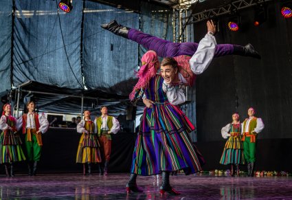 Na pierwszym planie para Artystów w kostiumach opoczyńskich , gdzie Artystka podnosi Artystę i wykonują spektakularne figury taneczne na scenie