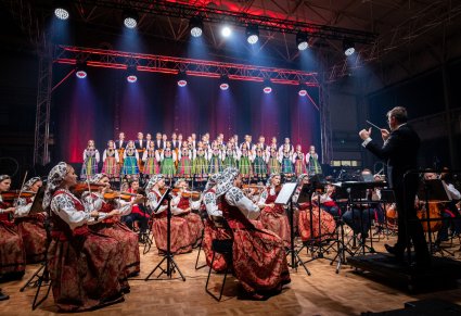 Dyrygent z batutą skierowany w stronę orkiestry i chóru w kostiumach łowickich