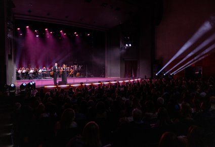 Na pierwszym planie na scenie para prowadzących koncert, na drugim planie Orkiestra