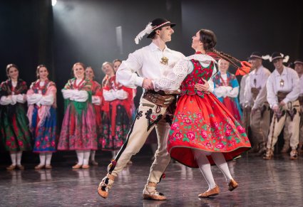 Na pierwszym planie para Artystów w kostiumach podhalańskich w trakcie wykonywania choreografii tanecznej, w tle reszta część Artystów