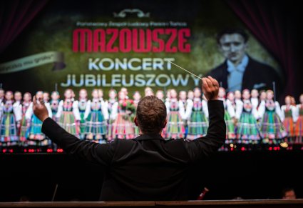 Na pierwszym planie Dyrygent z uniesioną batutą zwrócony w stronę orkiestry i Artystów na scenie