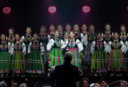 Na pierwszym planie Dyrygent zwrócony w stronę orkiestry i Chóru, na scenie na środku dwie Solistki a za nimi Artyści Chóru w kostiumach łowickich