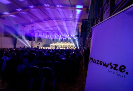 Na pierwszym planie logotyp Mazowsze Serce Polski a w tle widoczna publiczność z oświetloną sceną