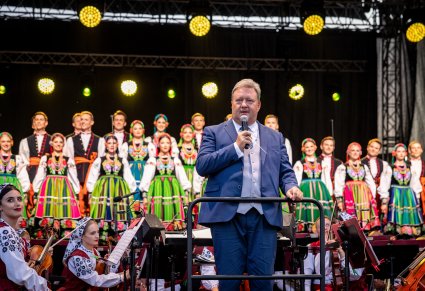 Dyrektor Jacek Boniecki wypowiadający się do mikrofonu, za nim orkiestra i chór na scenie w kostiumach łowickich