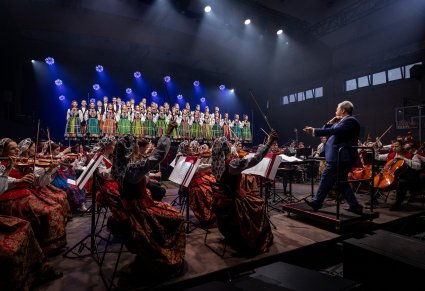 Na pierwszym planie Artyści Orkiestry wraz z Dyrygentem, trzymającym w ręku batutę, na scenie Artyści Chóru w kostiumach łowickich