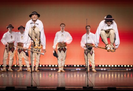 Na pierwszym planie dwóch Artystów w kostiumach podhalańskich w trakcie wykonywania spektakularnej choreografii skakania przez ciupagi, w tle część Artystów