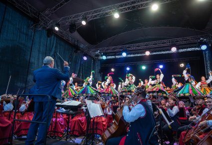 Na pierwszym planie Dyrektor Jacek Boniecki dyrygujący, w tle orkiestra i na scenie Artyści w ludowych kostiumach