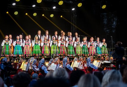 Chór w kostiumach łowickich i orkiestra na scenie z widoczną publicznością