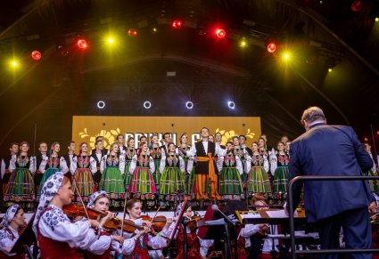 Dyrektor Jacek Boniecki zwrócony w stronę orkiestry i Artystów na scenie. Artyści z chóru mają na sobie kostiumy łowickie. Przed nimi stoi Solista