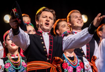 Zbliżenie na Solistę w trakcie śpiewu a za nim chór w kostiumach łowickich