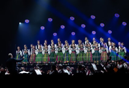 Na scenie Artyści Chóru w kostiumach łowickich, niżej sceny Dyrygent zwrócony w stronę Chóru i Orkiestry, trzymający w ręku batutę