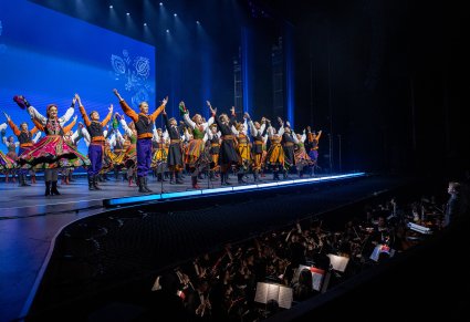 Ujęcie boczne na scenę na której są Artyści w kostiumach ludowych podczas wykonywania finałowej choreografii. Widoczny również orkiestron z Muzykami