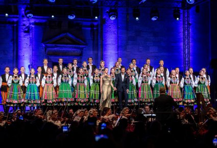 Na pierwszym planie Solistka w długiej, złotej sukni i obok niej Solista w eleganckim garniturze, przed nimi Orkiestra a za nimi Artyści z chóru w kostiumach łowickich