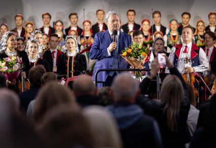 Widok z perspektywy publiczności na przemawiającego Dyrektora zespołu, trzymającego bukiet kwiatów, w tle orkiestra w kostiumach wilanowskich i chór w kostiumach łowickich