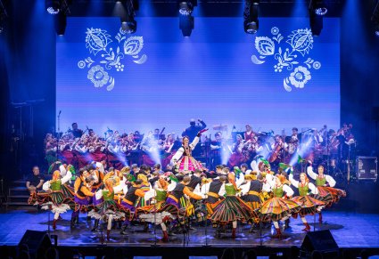 Na pierwszym planie na scenie Artyści w kostiumach ludowych podczas wykonywania choreografii tanecznej, w tle sceny Artyści Orkiestry w kostiumach wilanowskich wraz z Dyrygentem