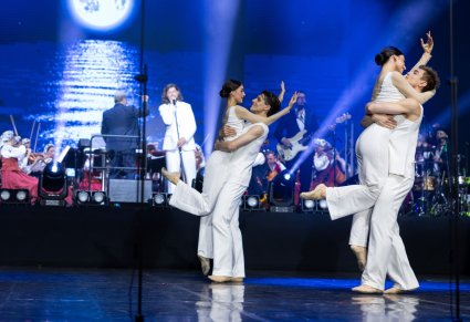 Na pierwszym planie na scenie pary Artystów w kostiumach współczesnych w trakcie wykonywania choreografii tanecznej, w tle widoczna część Orkiestry w kostiumach wilanowskich wraz z Dyrygentem