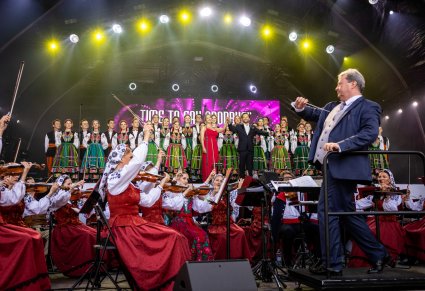 Dyrektor Jacek Boniecki dyrygujący koncert, zwrócony jest w stronę orkiestry. Na scenie dwoje Solistów, kobieta w czerwonej sukni i mężczyzna w garniturze a za nimi Artyści chóru w kostiumach łowickich