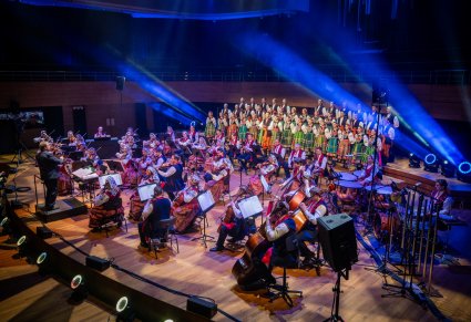 Widok na scenę na której znajduje się Dyrygent, Orkiestra w kostiumach wilanowskich i Artyści Chóru w kostiumach łowickich