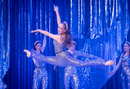 Na pierwszym planie skacząca szpagat w górze postać Gwiazdy w srebrnym kostiumie