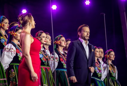 Zbliżenie na parę Solistów. Kobieta ubrana w czerwoną suknię, mężczyzna w ciemny garnitur. Za nimi część Artystów z chóru