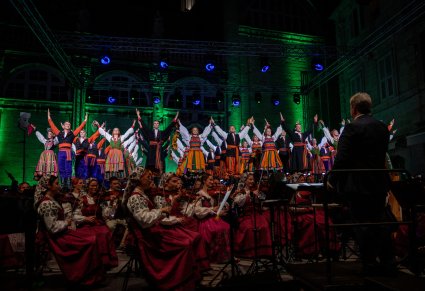 Artyści w różnych kostiumach ludowych w trakcie wykonywania finałowego układu tanecznego a przed nimi orkiestra z Dyrektorem Jackiem Bonieckim, Dyrygentem koncertu