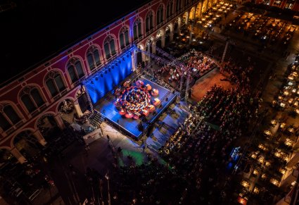 Widok nocny na koncert plenerowy w otwartym dziedzińcu,  z publicznością, sceną oświetloną kolorowymi światłami