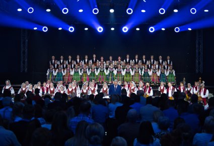 Widok na scenę na której znajdują się Artyści Chóru w kostiumach łowickich, Artyści Orkiestry w kostiumach wilanowskich oraz Dyrygent, widoczna również część Publiczności
