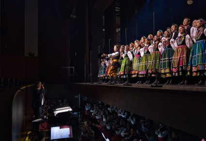 Widok na Artystów Chóru w kostiumach łowickich, stojących na scenie oraz na Orkiestron w którym znajduje się Dyrygent, trzymający w ręku batutę oraz Artyści Orkiestry w kostiumach wilanowskich