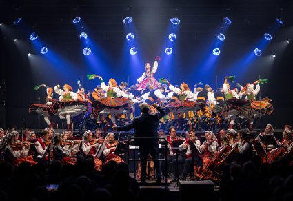 Na scenie Artyści w kostiumach ludowych w rakcie wykonywania choreografii tanecznej, niżej sceny Artyści orkiestry w kostiumach wilanowskich wraz z Dyrygentem trzymającym w ręku batutę