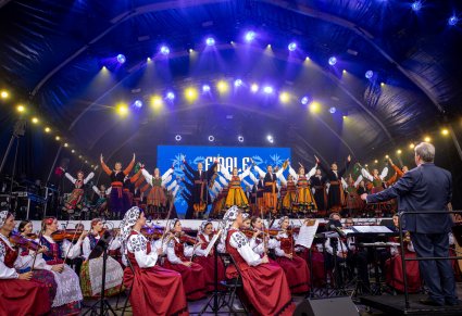 Dyrektor Jacek Boniecki zwrócony w stronę Artystów. Orkiestra znajduje się niżej od podestu z Artystami wykonującymi choreografię finałową. Artyści ubrani są w kostiumy ludowe