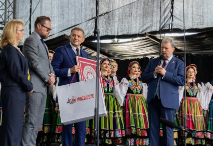 Na scenie mer rejonu solecznickiego wraz z przedstawicielami Samorządu Województwa Mazowieckiego i Dyrektor zespołu "Mazowsze" w tle część zespołu w kostiumach łowickich