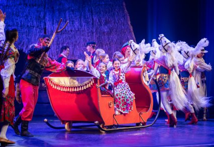 Dziewczynki grające w spektaklu siedzące w czerwonych saniach, które ciągnięte są przez postacie koni. Obok nich postać diabła ludowego i góralki