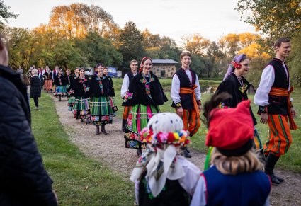 Artyści w kostiumach łowickich i wilanowskich, po koncercie, idący wśród publicznosći, obserwowani przez parę dzieci w kostiumach