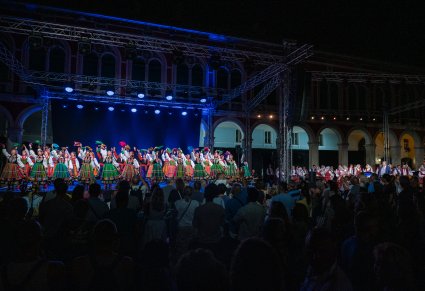 Grupa tancerzy w kostiumach łowickich występuje na oświetlonej scenie podczas nocnego koncertu plenerowego przed publicznością.