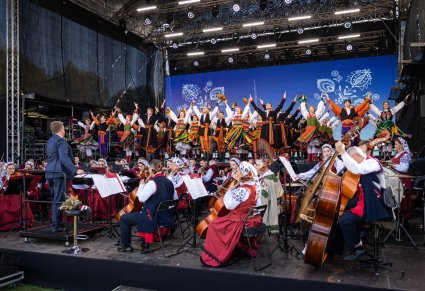Widok na scenę na której znajduje się Dyrygent z batutą, Orkiestra w kostiumach wilanowskich i Artyści w kostiumach ludowych, którzy wykorzystują choreografię taneczną