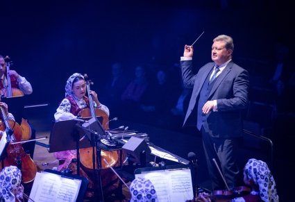 Zbliżenie na Dyrektora zespołu "Mazowsze", który trzyma w ręku batutę, przed nim część Orkiestry w kostiumach wilanowskich