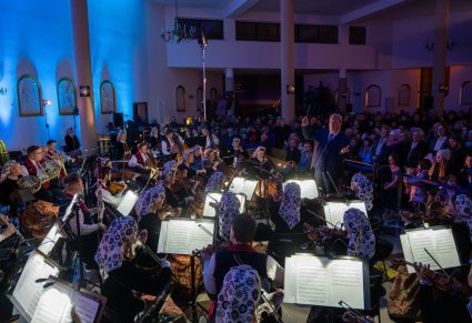 Na pierwszym planie Dyrektor zespołu w trakcie dyrygowania, przed nim Orkiestra w kostiumach wilanowskich, za nim publiczność koncertu kolęd i pastorałek