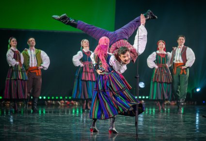 Na pierwszym planie para Artystów  w kostiumach opoczyńskich podczas wykonywania imponujących figur tanecznych, za nimi w tle reszta część Artystów również w kostiumach opoczyńskich