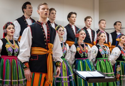 Na pierwszym planie Solista Chóru w kostiumie łowickim, w tle reszta część Chóru w trakcie śpiewu, również w kostiumach łowickich