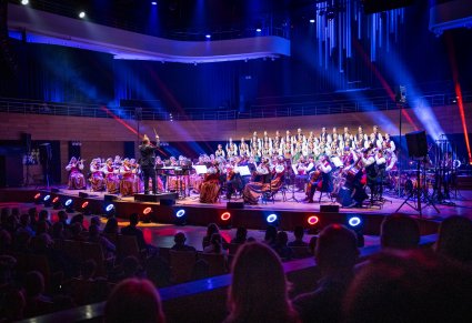 Widok na scenę na której znajduje się Dyrygent zwrócony w stronę Orkiestry w kostiumach wilanowskich i Chór w kostiumach łowickich