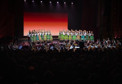 Szeroki widok na scenę i publiczność. Na scenie Artyści Chóru w kostiumach łowickich w trakcie śpiewu,  niżej sceny Artyści Orkiestry w kostiumach wilanowskich i Dyrygent