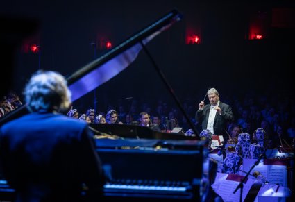 Na pierwszym planie Dyrygent zwrócony w stronę pianisty przy fortepianie, widoczna Orkiestra w kostiumach wilanowskich