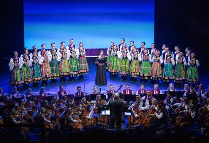 Na środku sceny Solistka w długiej, czarnej sukni, po jej bokach Artyści Chóru w kostiumach łowickich, niżej sceny Artyści Orkiestry w kostiumach wilanowskich wraz z Dyrygentem