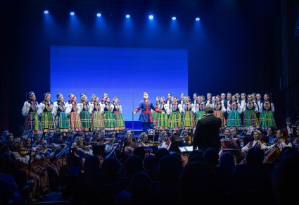 Na scenie Artyści Chóru w kostiumach łowickich a na środku sceny Solista w kostiumie kujawskich w trakcie śpiewu. Widoczna Orkiestra w kostiumach wilanowskich wraz z Dyrygentem