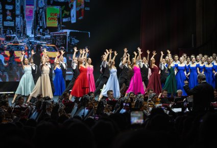 Na scenie Artyści Baletu w kolorowych współczesnych kostiumach w trakcie wykonywania choreografii tanecznej, z boku sceny Artyści chóru we współczesnych strojach podczas śpiewu, niżej sceny Artyści Orkiestry w kostiumach wilanowskich wraz z Dyrygentem